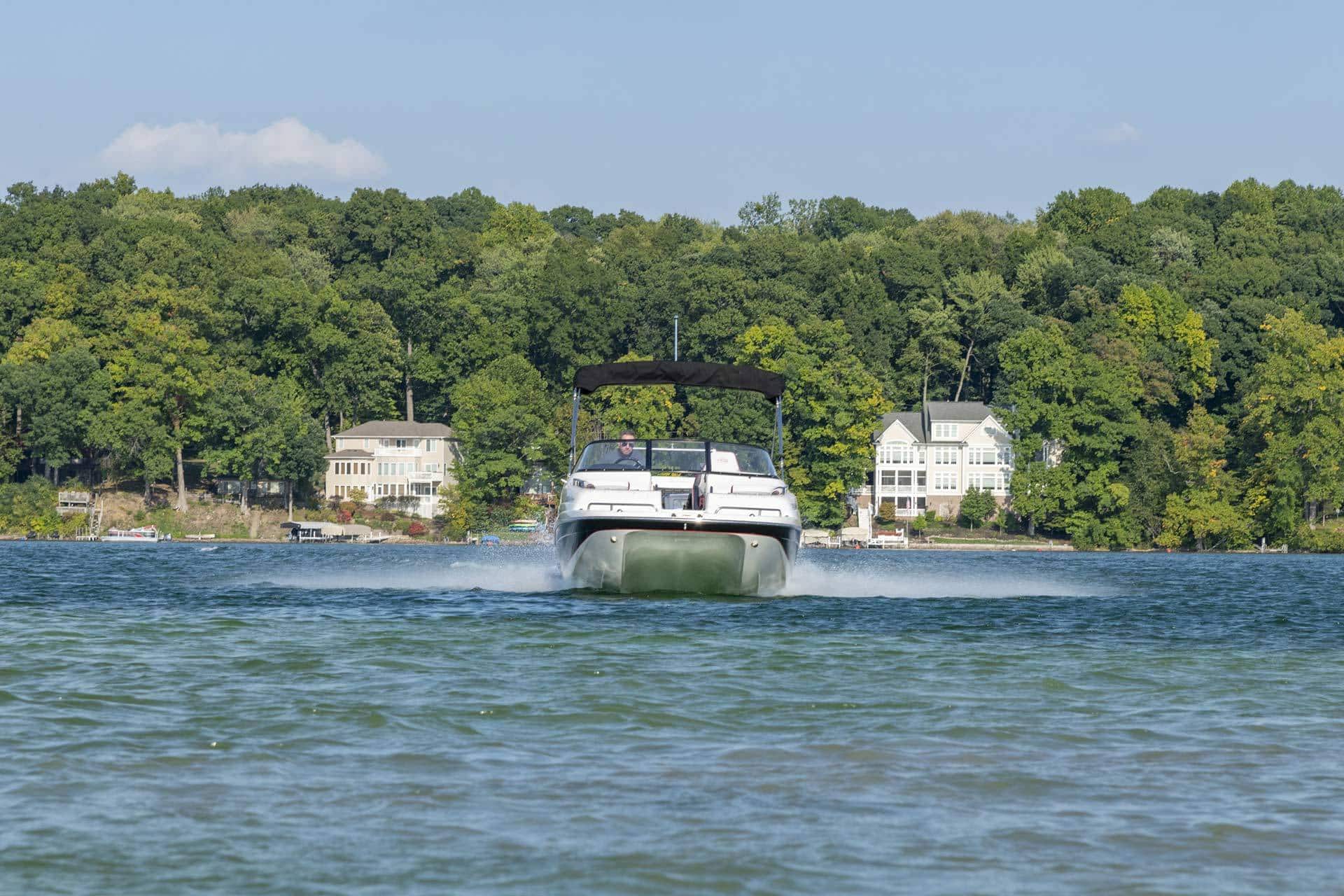 Man driving a Splendor Boats catamaran boat on a lake with large houses and trees in the background.