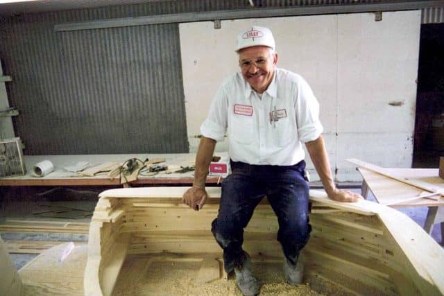 A smiling Caucasian man in a white hat, white shirt, and blue pants sitting on a partially constructed Splendor deck boat.